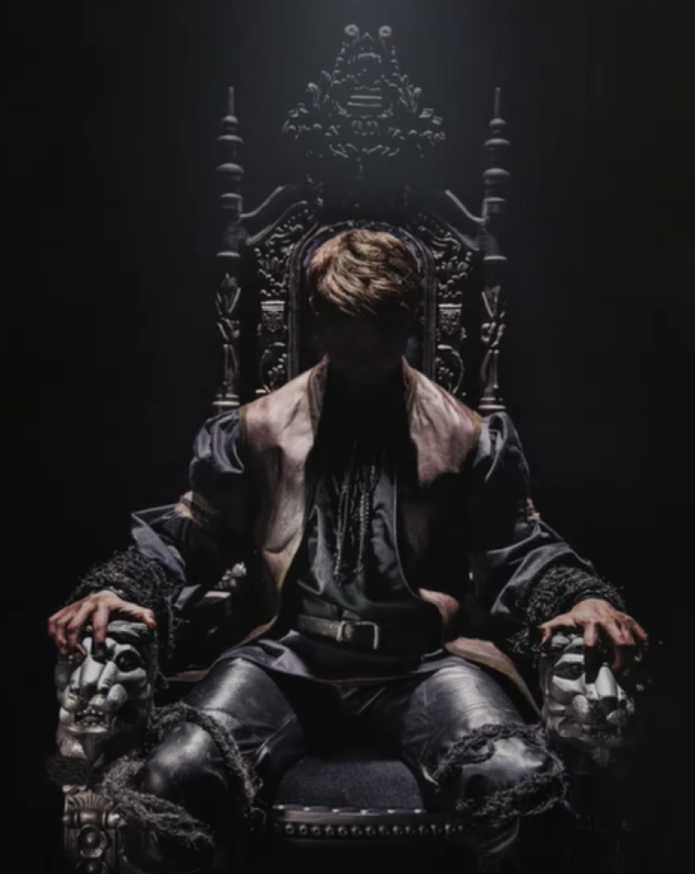 A person with brown hair is sitting on an ornate, dark wooden throne with a high back. The person's face is obscured in shadow, highlighting only the silhouette. They are dressed in dark, layered clothing, and their hands rest on carved skulls that a
