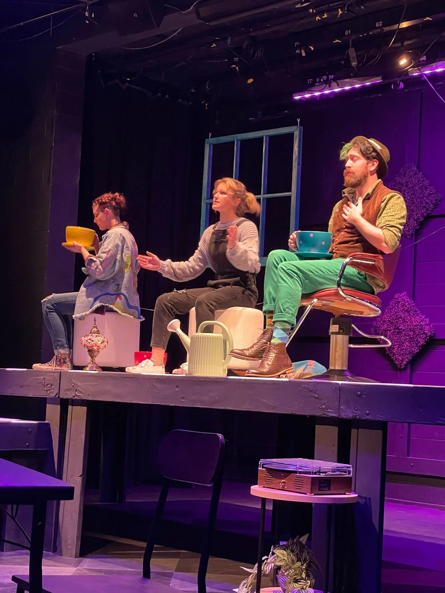 A stage with three actors seated with props such as bowls, a watering can, and toys, part of a theatrical performance. Erika McGonigal Johnson plays Alice in “Back to Wonderland” by Lily Fonte.
