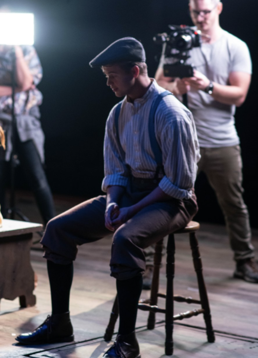 A young man dressed in historical costume, sitting on a wooden stool during a film or theater production, with a crew member operating a camera in the background, illuminated by studio lights. Jed Johnson Plays Gilbert Blithe in Anne of Green Gables.