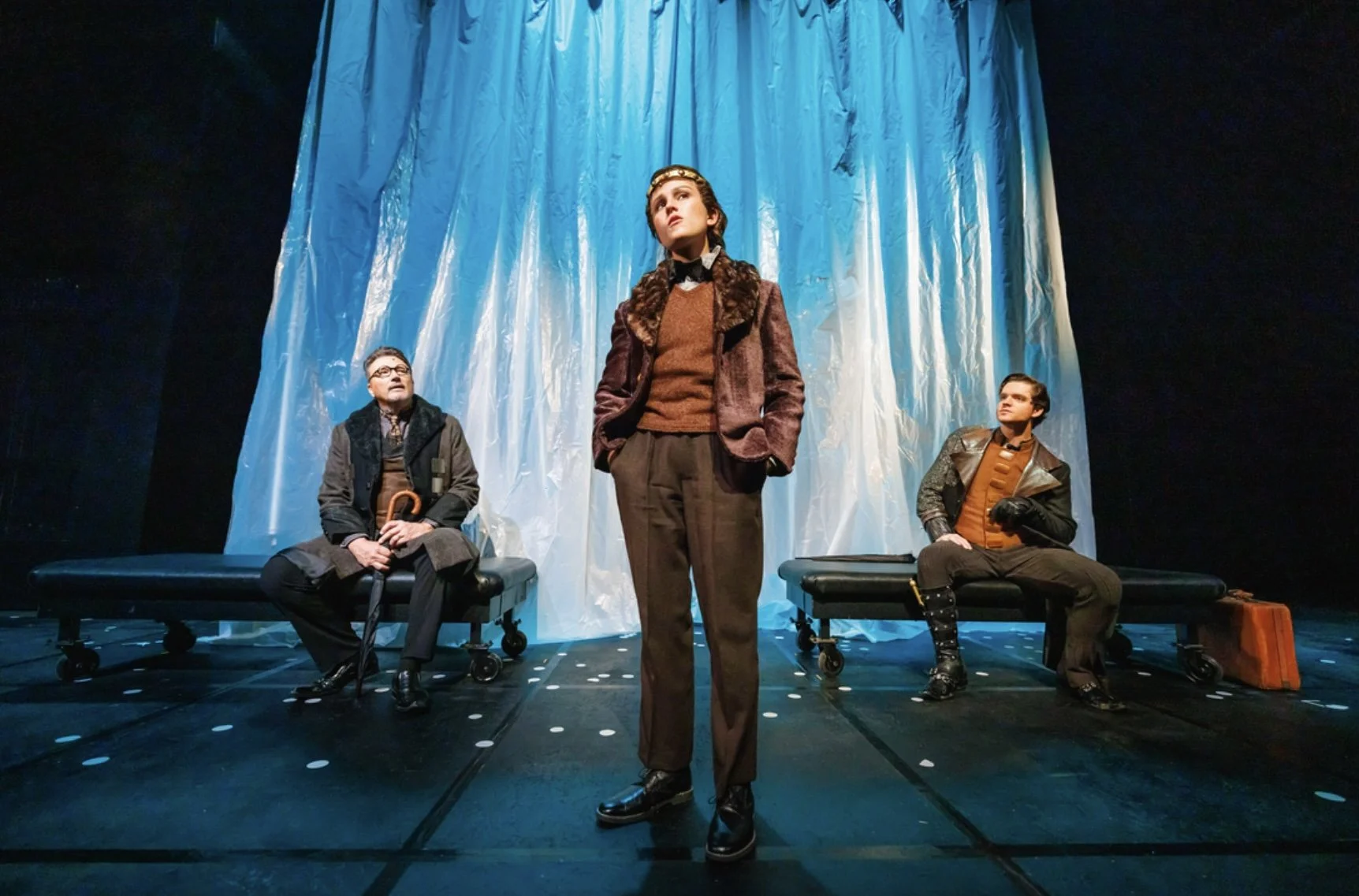 Three actors on a stage, with a woman standing in front and two men sitting on either side. The woman is wearing a brown jacket and brown trousers, looking contemplative. The man on the left is dressed in darker, vintage clothing with glasses and a c