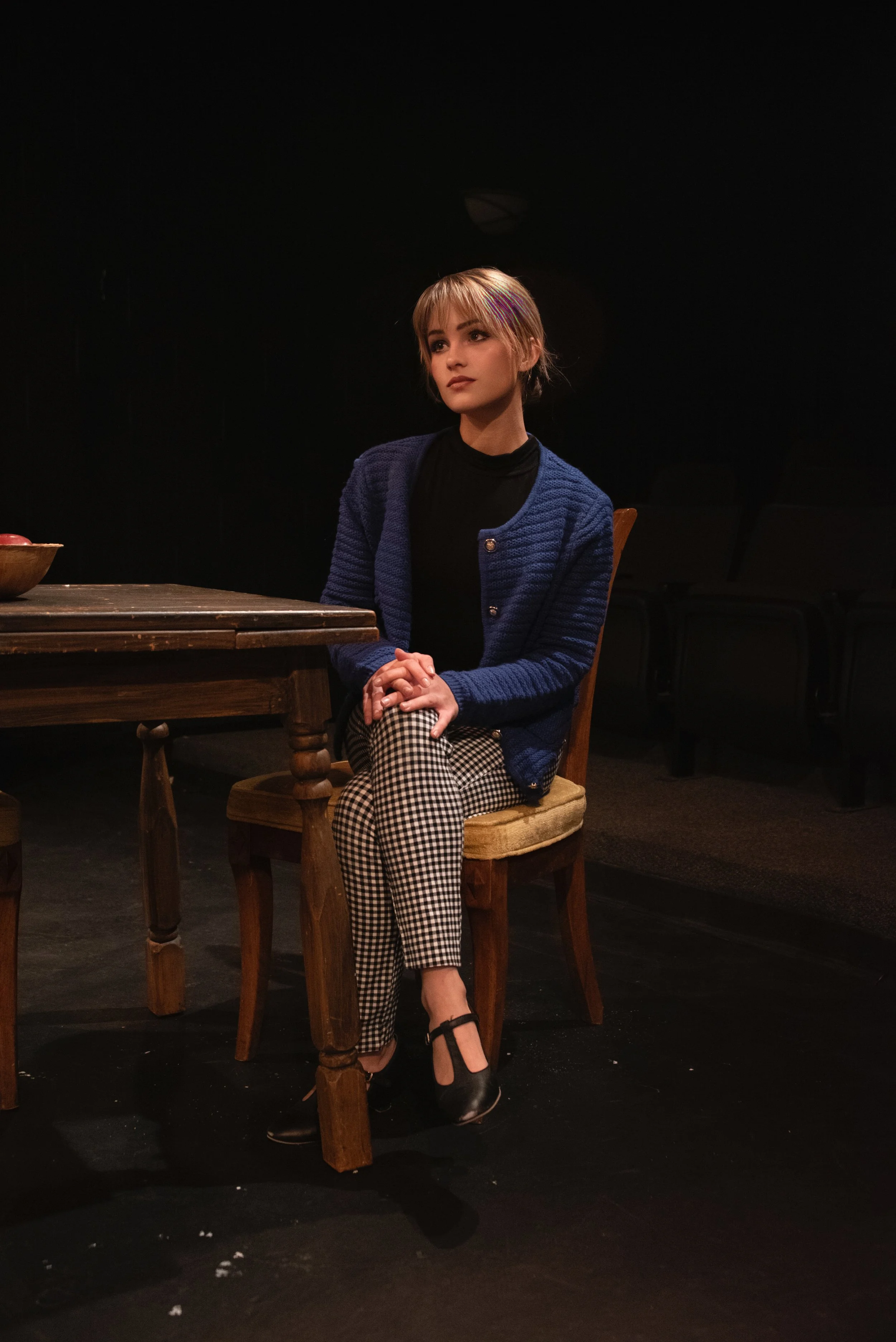 A woman with short blonde hair sitting at a wooden table with her hands clasped, wearing a blue cardigan and black-and-white checkered pants, in a dimly lit setting.  Erika McGonigal Johnson plays Susy Hendrix in “Wait Until Dark.”