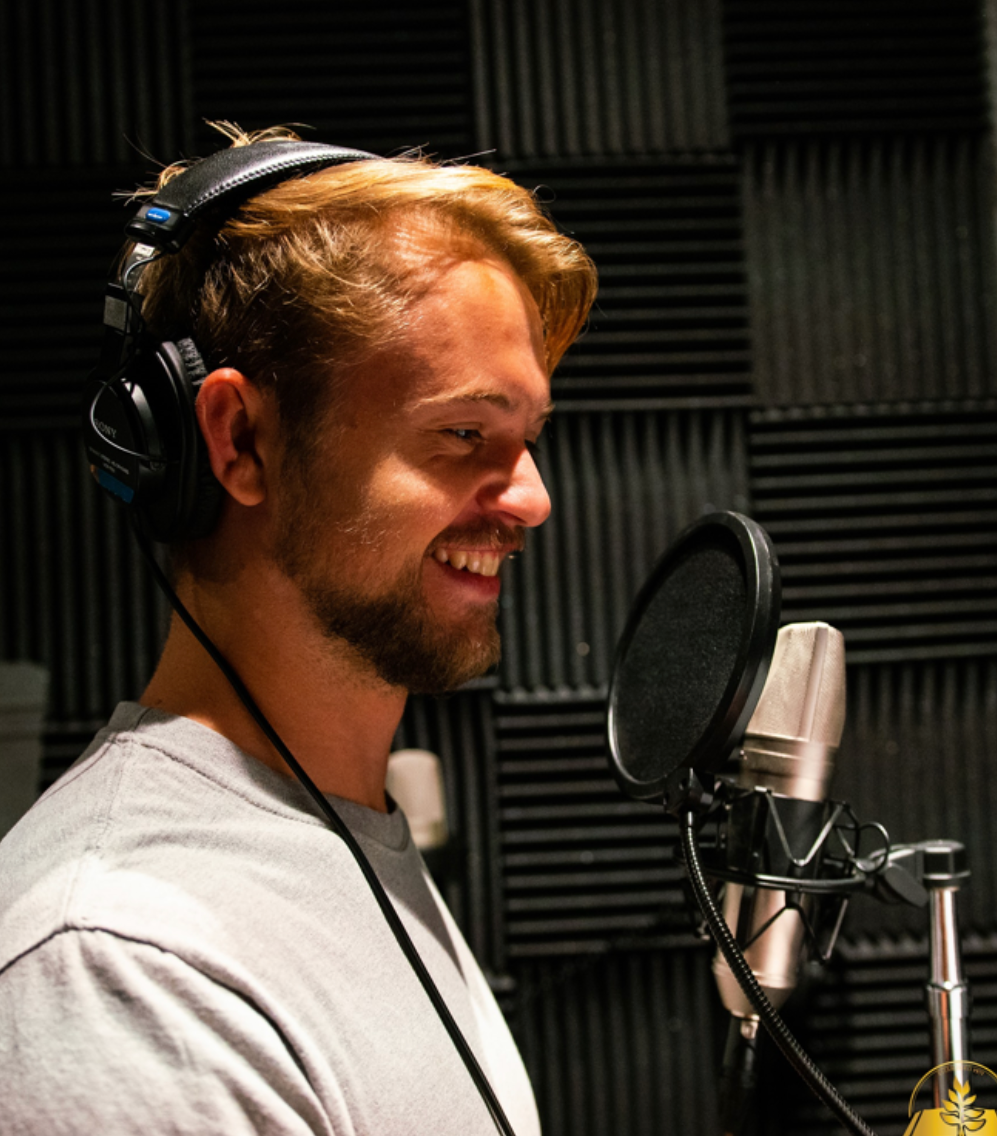 A young man with light brown hair, a beard, and wearing headphones, is smiling and standing in front of a professional microphone in a recording studio with soundproof foam panels.