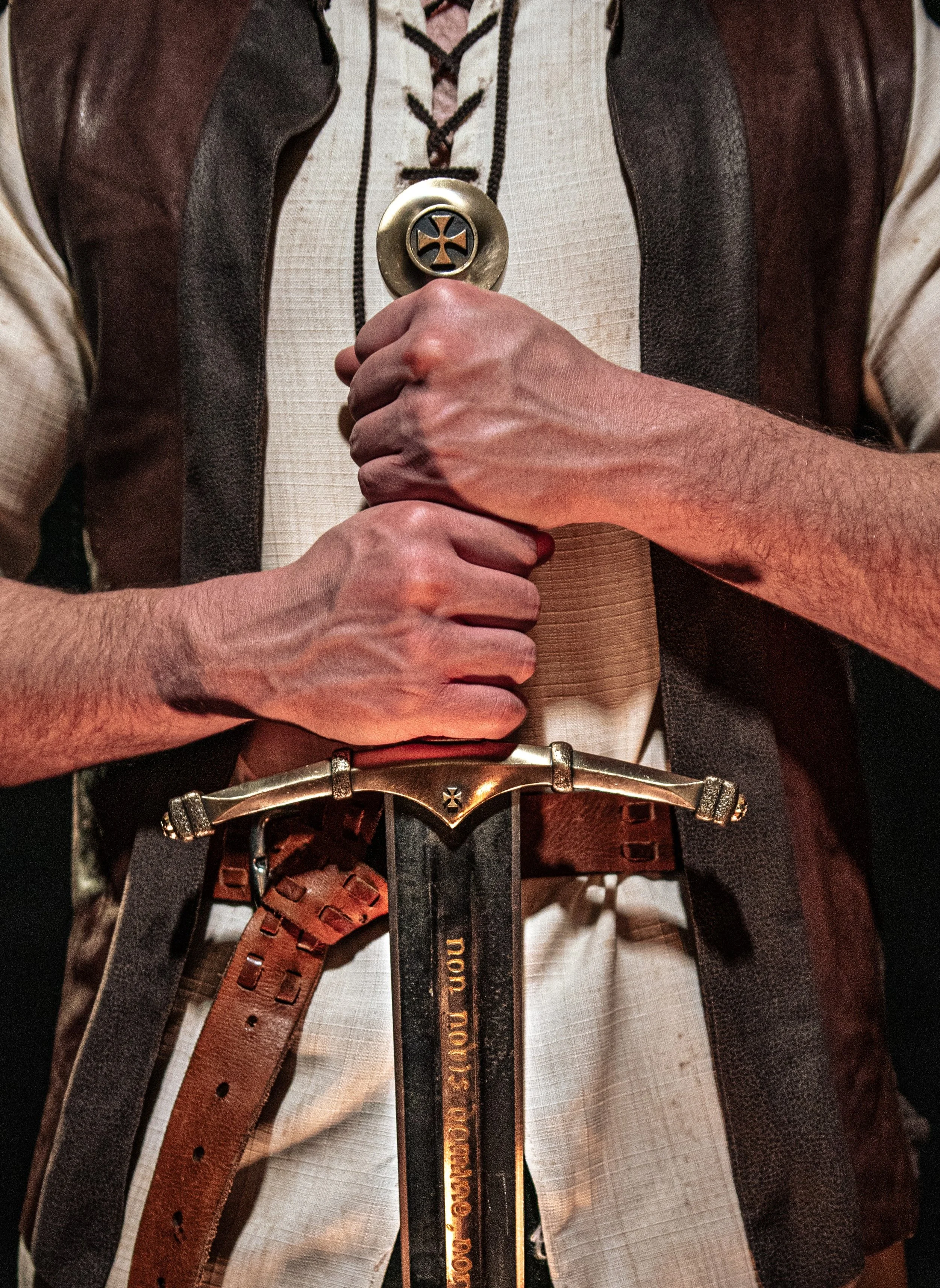 Person wearing a leather vest over a white shirt, holding a large metallic cross pendant necklace with a circular frame. They have a brown leather strap belt with metal accents. The photo focuses on the chest and hands.