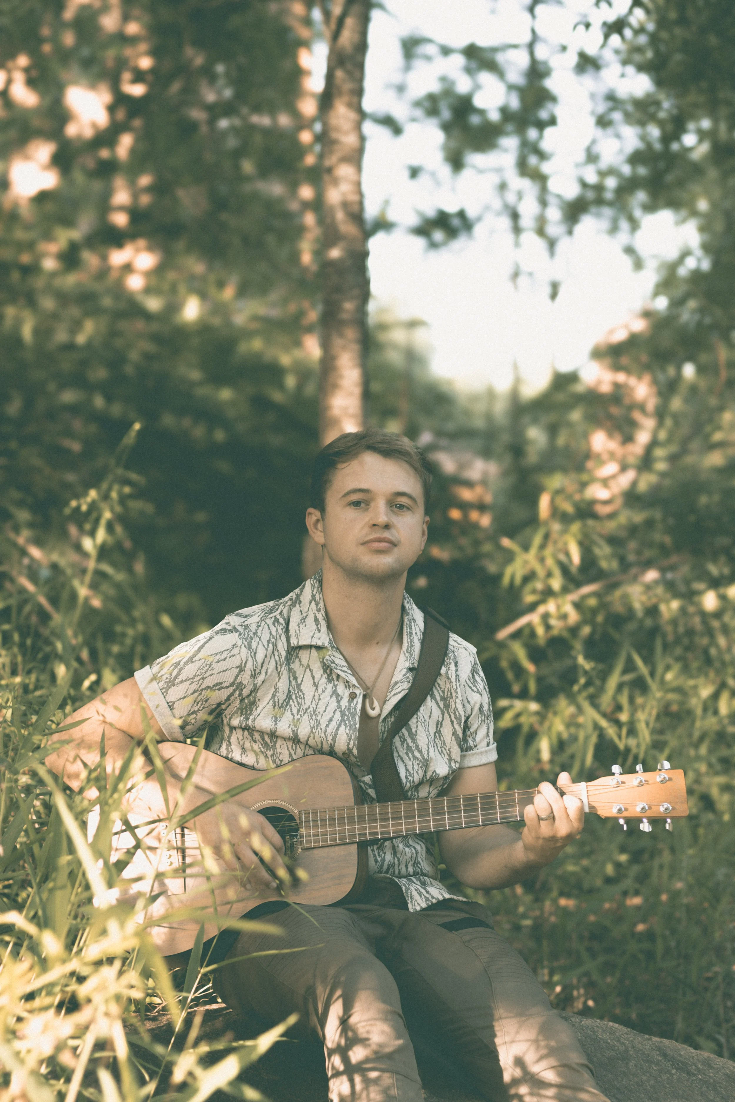 Jed Johnson, a young man sitting outdoors among greenery with trees in the background, playing an acoustic guitar and looking into the distance in the late afternoon.