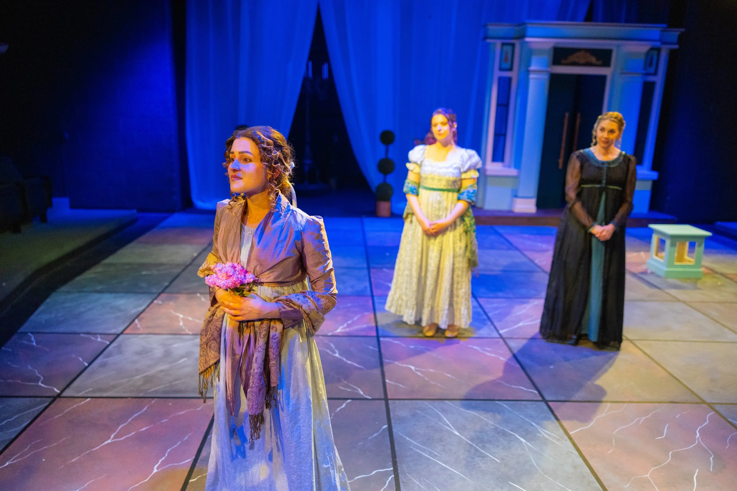 Three women in historical costumes standing on a stage with a blue background and curtains. Erika McGonigal Johnson plays Marianne Dashwood in Sense and Sensibility BJU 2025.