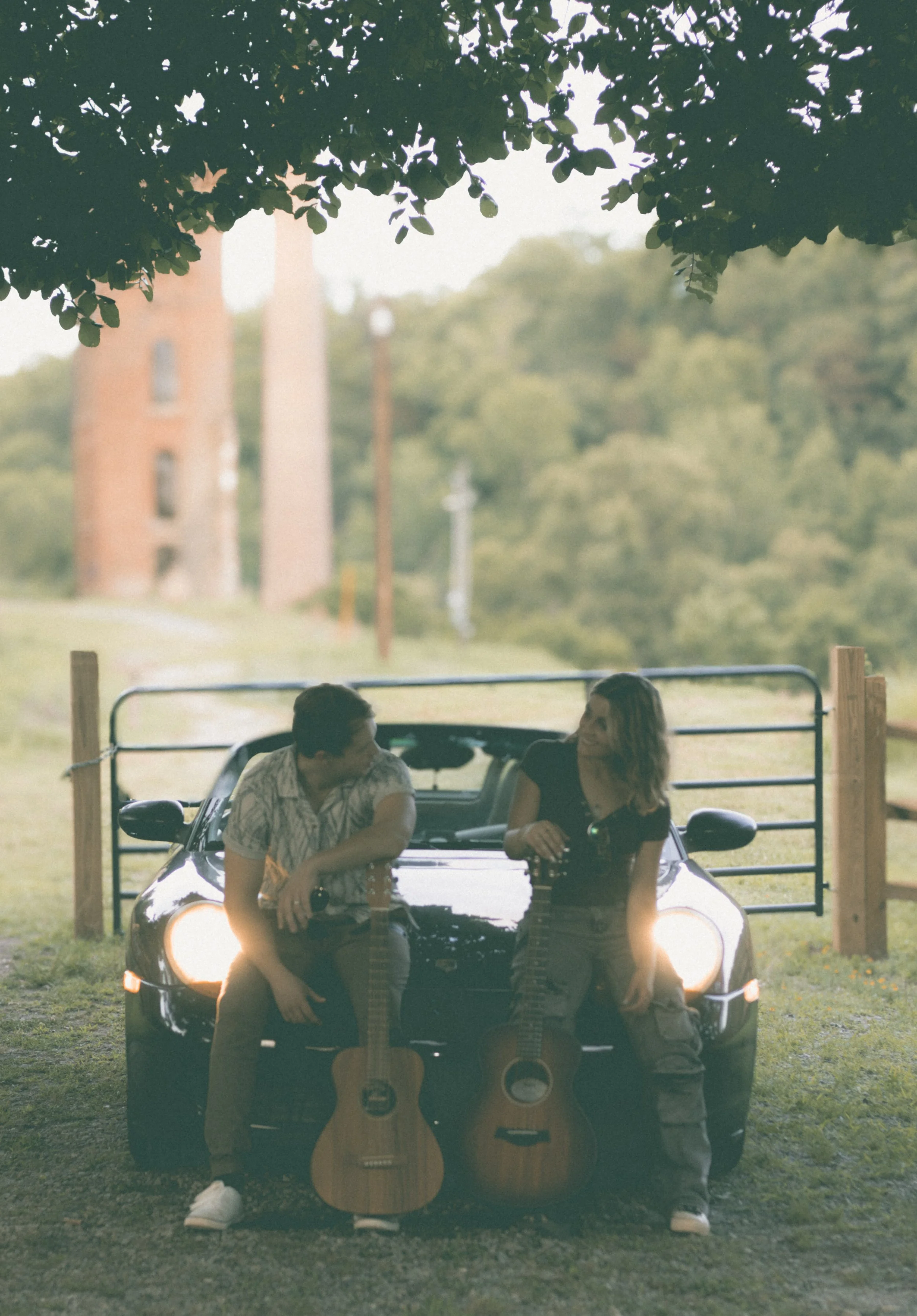 Two people sitting on a car with guitars hanging on the front, in an outdoor setting with trees and a fence, during daylight.