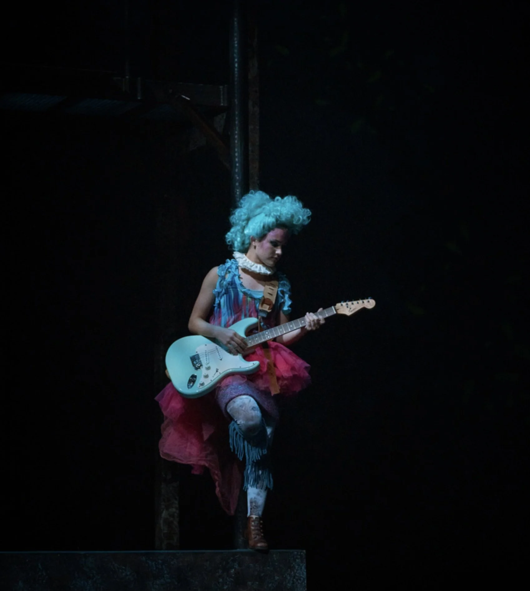 Person dressed in a colorful costume with a curly teal wig, playing an electric guitar on dark stage.