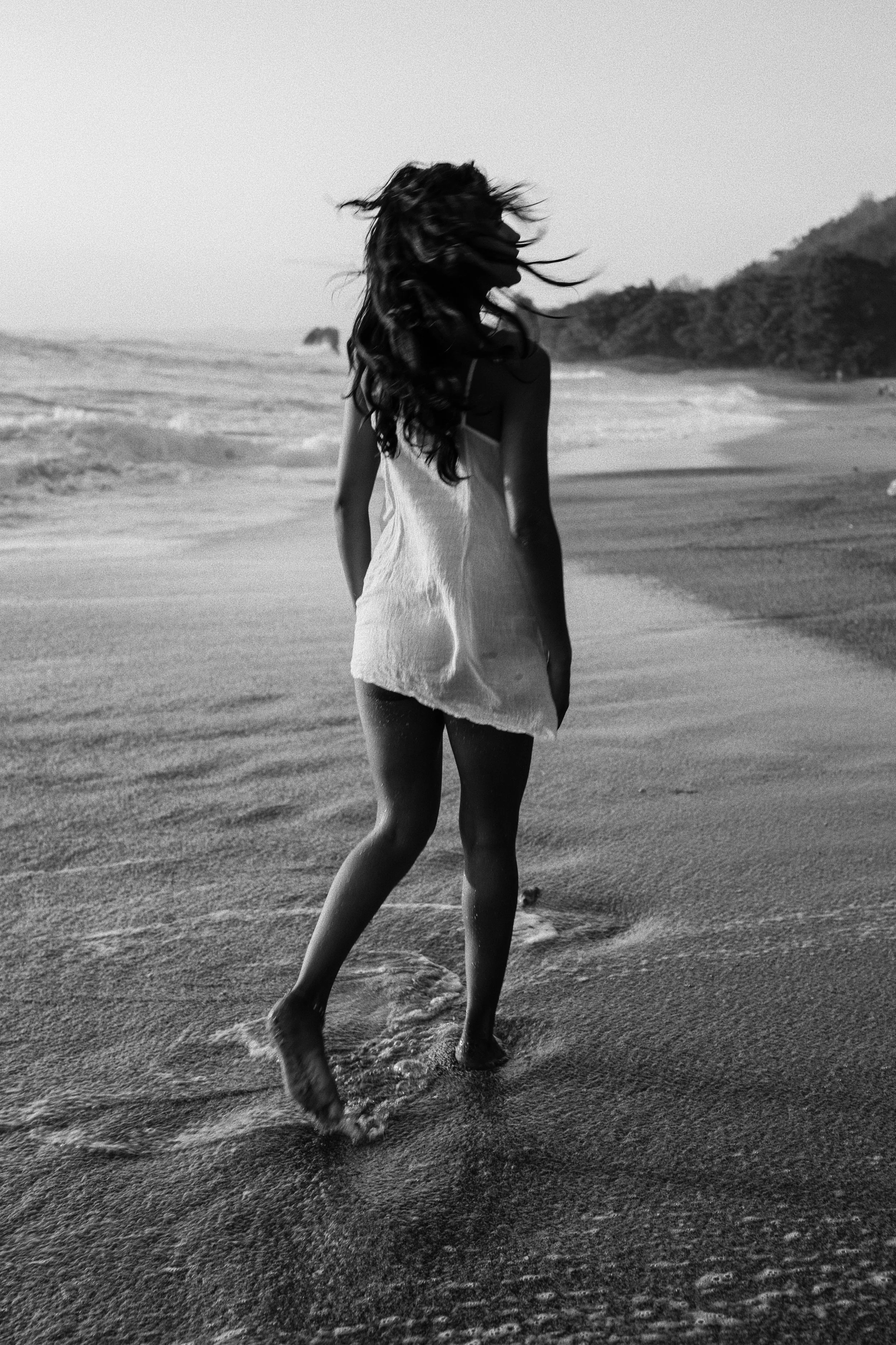 A woman with long, curly hair walks along a beach in black and white, with hills in the background and ocean waves nearby.