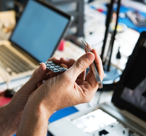 Person holding a small electronic device and a stylus pen in an electronics workshop or repair shop with laptops and tools in the background.