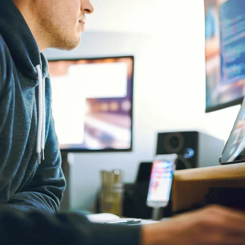 A person sitting at a desk with a computer monitor, a smartphone, and a speaker, working or browsing.