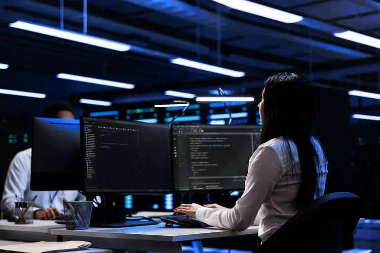 A woman working on multiple computer monitors in a dark, high-tech office environment.