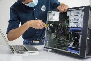 A person wearing a mask working on the inside of a desktop computer with a screwdriver, next to a laptop on a table.