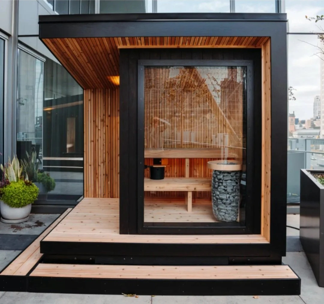 A modern rooftop sauna with black exterior panels and wooden interior, featuring a glass door and a small deck area outside.