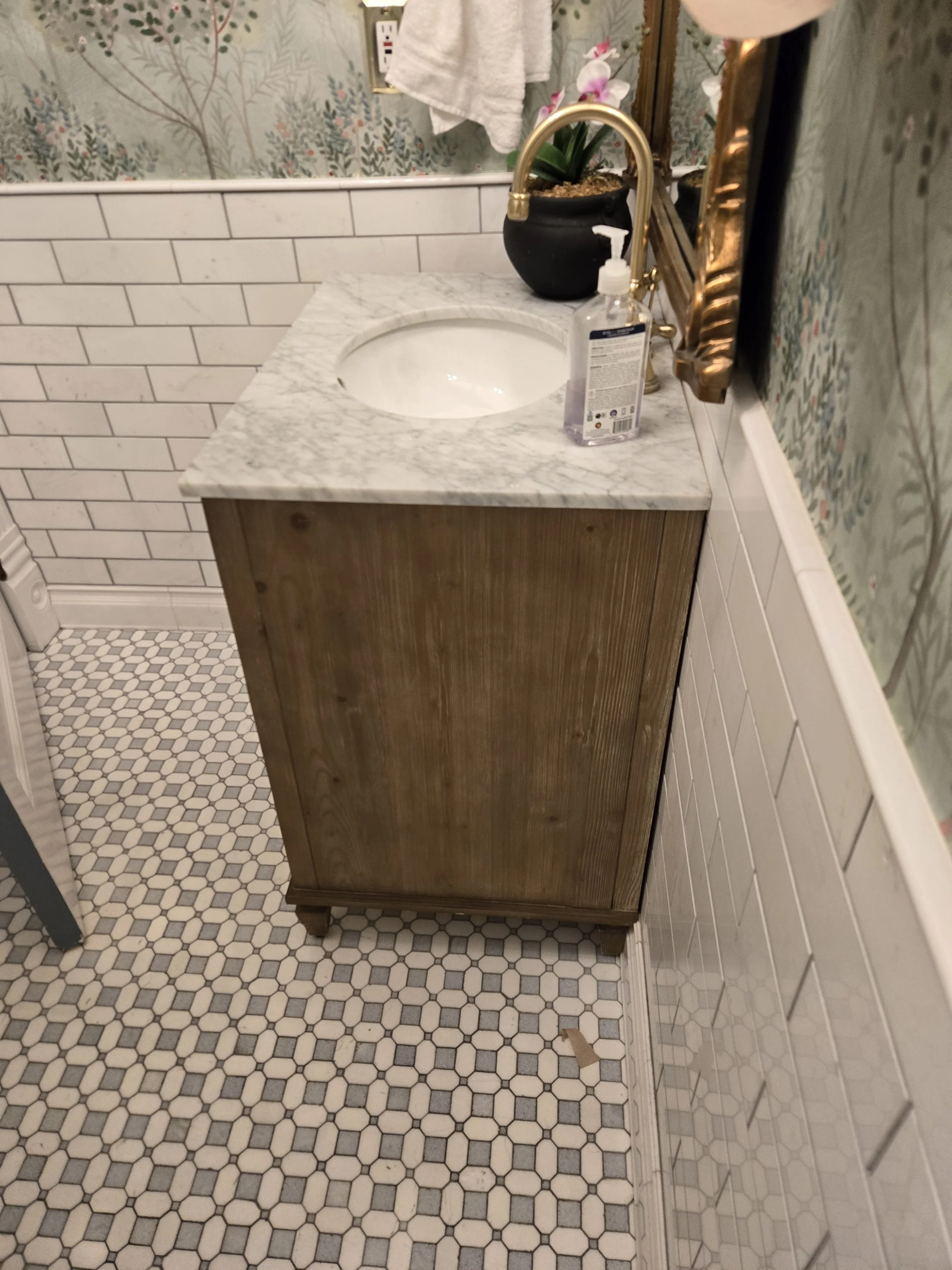 Bathroom vanity with a marble countertop, a sink, a gold faucet, a potted orchid plant, a bottle of hand soap, a towel hanging on the wall, and patterned tile flooring.