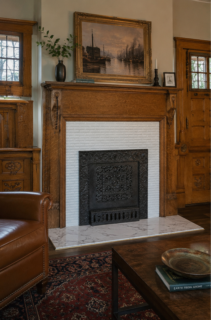 A cozy living room featuring a white brick fireplace with an ornate black metal screen. Above the fireplace hangs a framed painting of a harbor scene with ships and smoke. To the left, a dark vase with greenery sits on the mantle, next to books and a