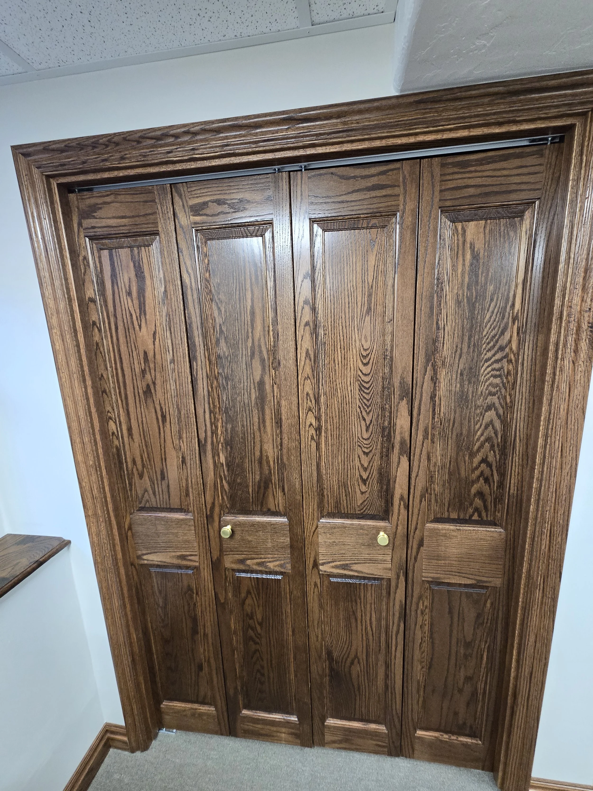 A wooden closet with six paneled doors, featuring brass knobs, surrounded by wooden trim and molding, situated against a white wall and carped floor.