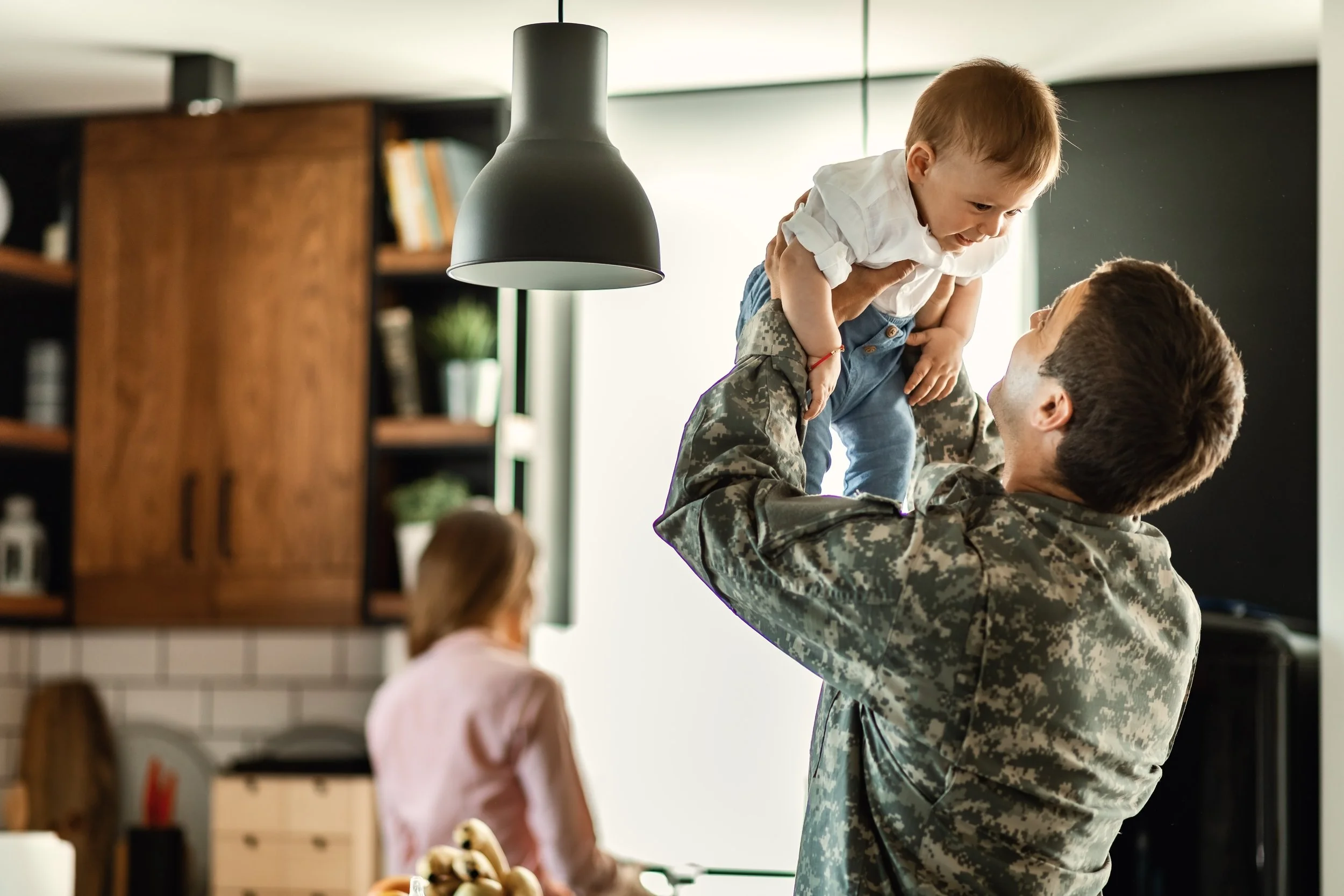 A person in a camouflage military uniform lifting a smiling young boy in a white shirt in a bright room with shelves and books.