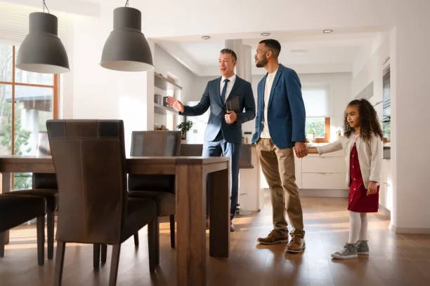 A family of three, two men and a young girl, holding hands inside a modern kitchen with dining area, bright natural light, and contemporary decor.