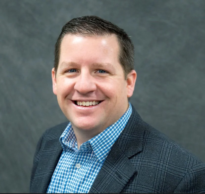 Headshot of a man in a checkered blue shirt and dark blazer smiling against a gray background.