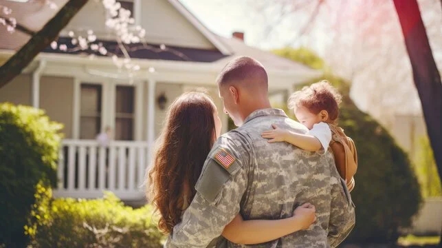 A soldier in uniform holding a young girl with curly hair outdoors, with a woman standing nearby. In the background, there is a house with a porch, trees, and blooming flowers.