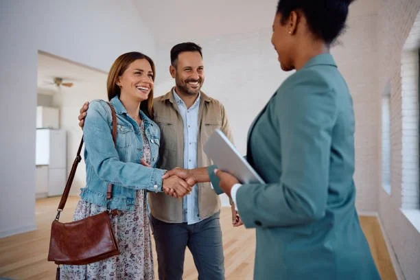 A woman in a denim jacket shaking hands with a real estate agent while a man stands beside her, smiling in a bright, modern home.