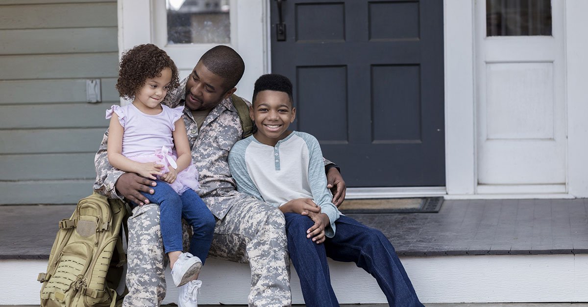 A soldier in uniform sitting on a porch with a young girl and boy, smiling and enjoying time together.