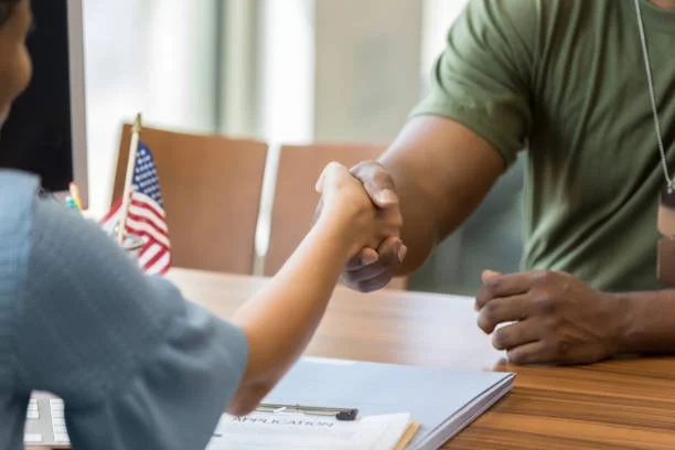 Person in a green shirt shaking hands with a child at a table, with American flags in the background.