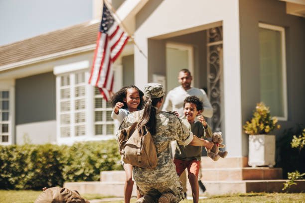 A soldier embracing two children, a girl and a boy, in front of a house with an American flag and a man standing nearby.