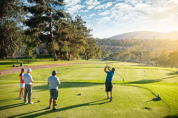 Four people playing golf on a lush green course, with a golfer swinging a club, and three others watching, surrounded by trees and mountains in the background during sunset.
