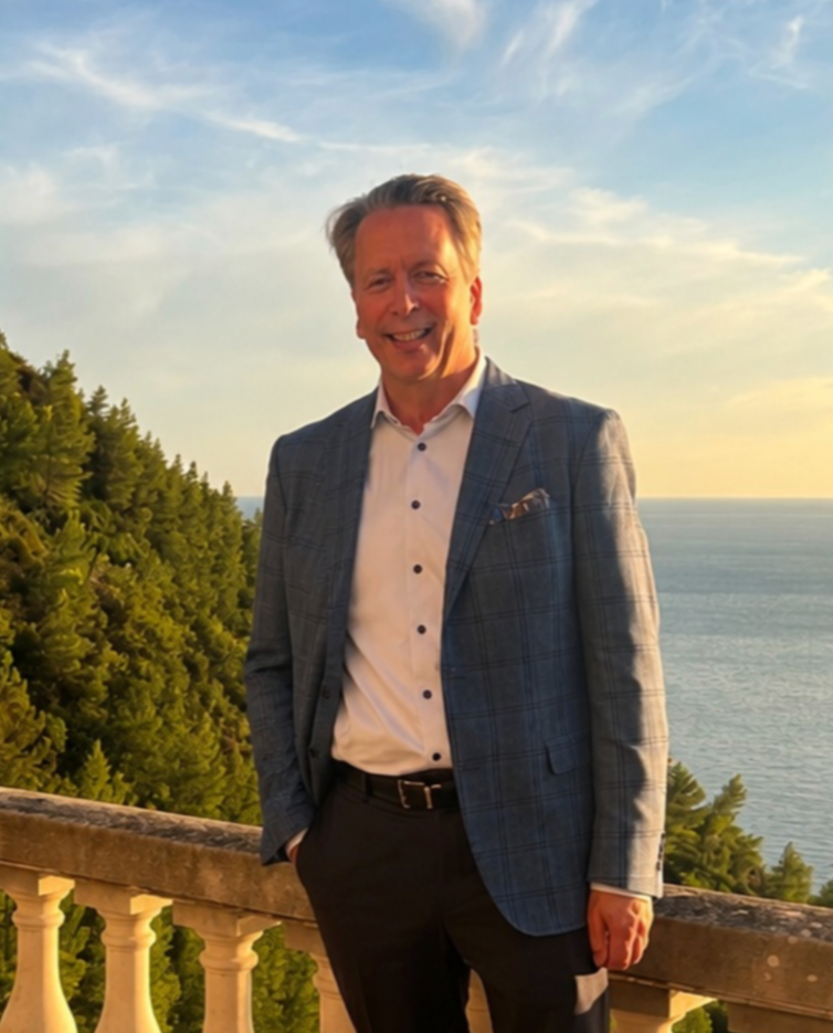 A man in a blue plaid suit jacket and white shirt, smiling outdoors on a balcony overlooking the ocean and green trees during sunset.