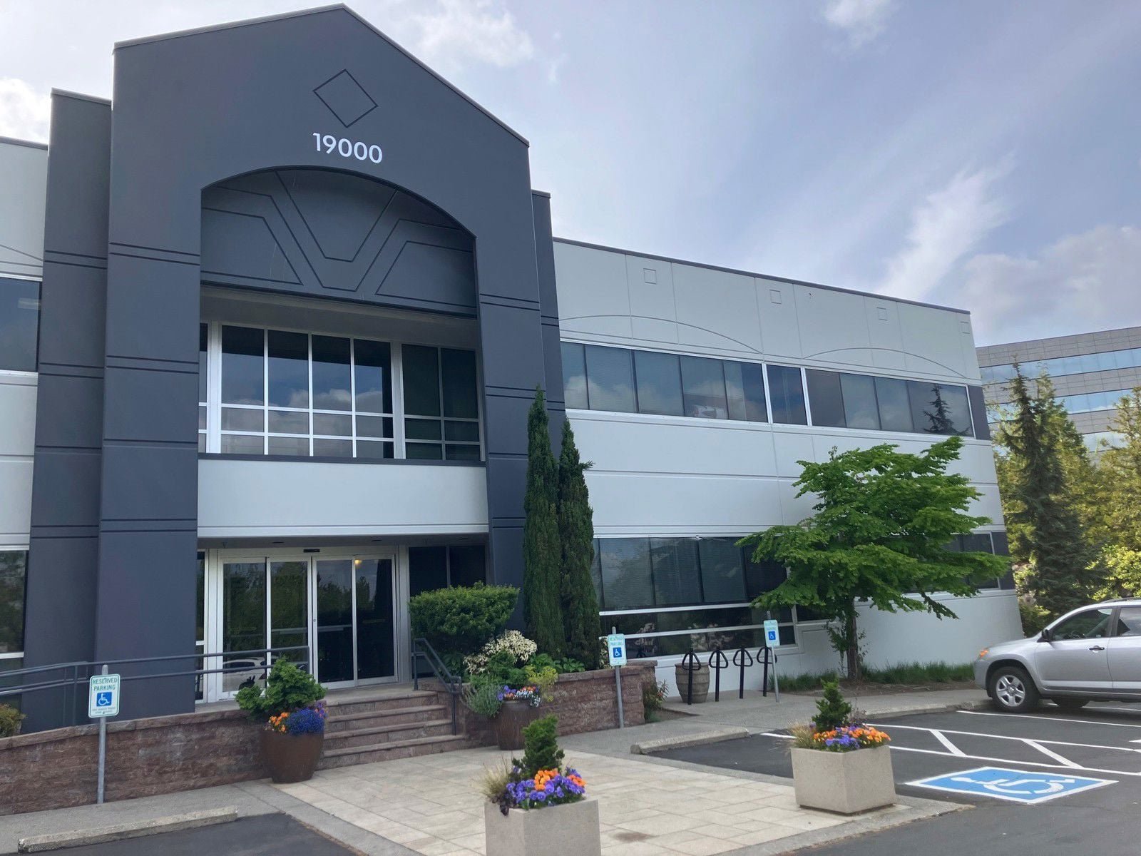 Exterior view of a modern office building with a parking lot, including designated accessible parking spaces, greenery, and flowers in pots.