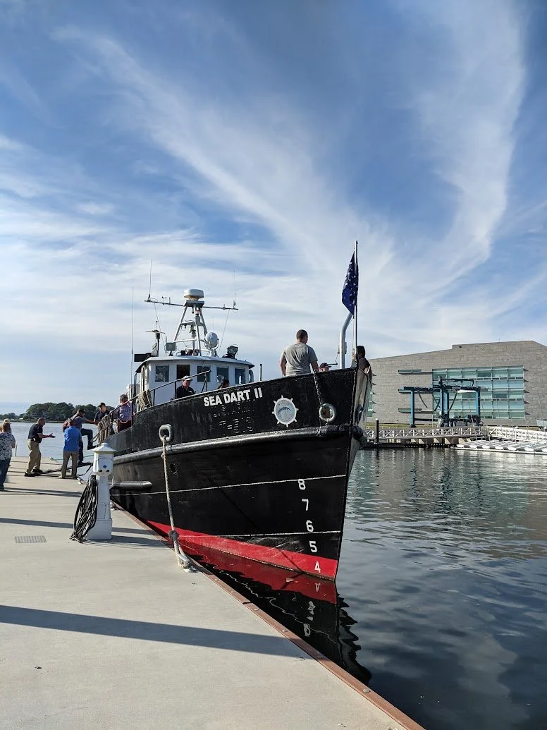 A black boat named 'Sea Dart II' docked at the pier with people on board and on the dock, with a modern building in the background and a blue sky with scattered clouds above.