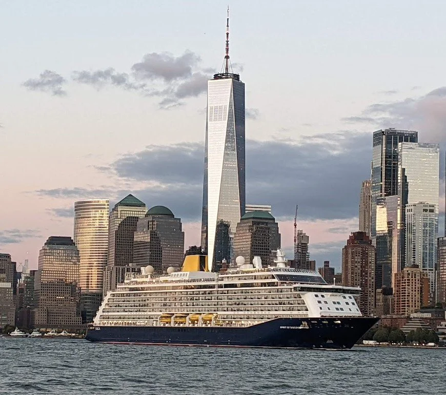 SAGA Holidays cruise ship sailing in front of a city skyline featuring One World Trade Center in New York City, with several surrounding tall buildings and gray clouds in the sky.
