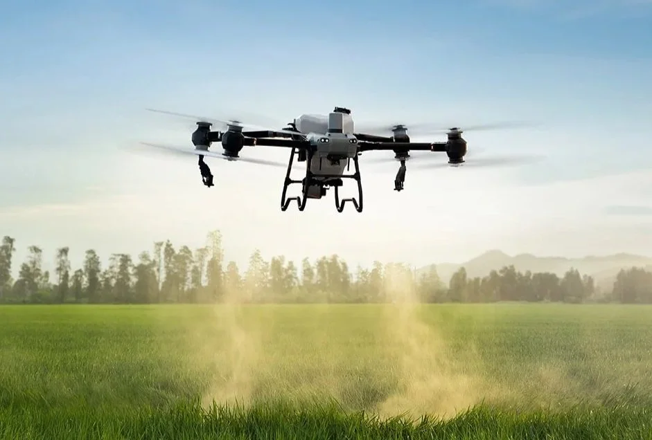 A drone flying over a green field emitting a dust or pesticide spray with trees and mountains in the background.