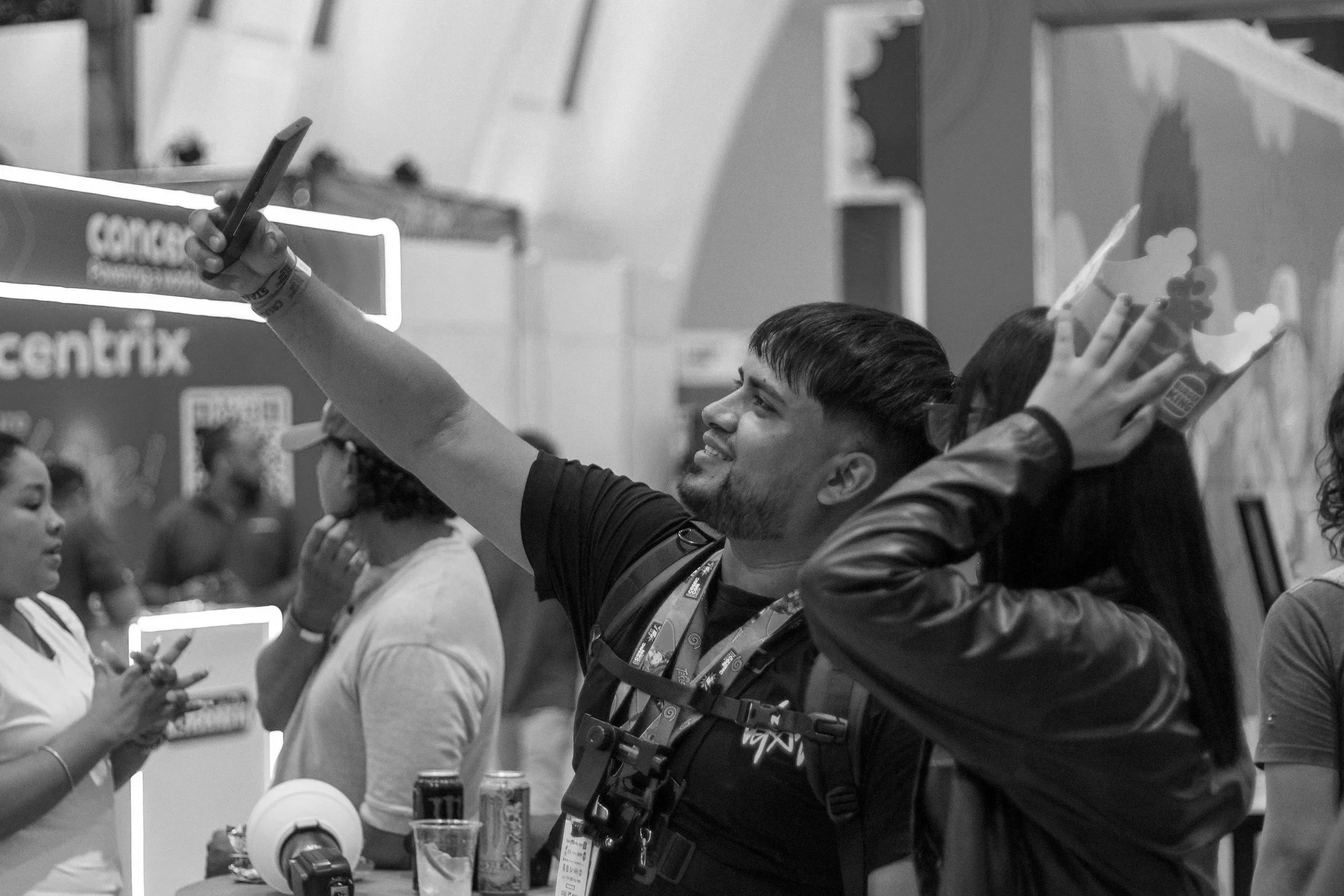 A man is taking a selfie at a table during an event, smiling and holding his phone up. Several other people are seen in the background, some engaging with their phones. The scene appears to be indoors with event booths and signage visible.