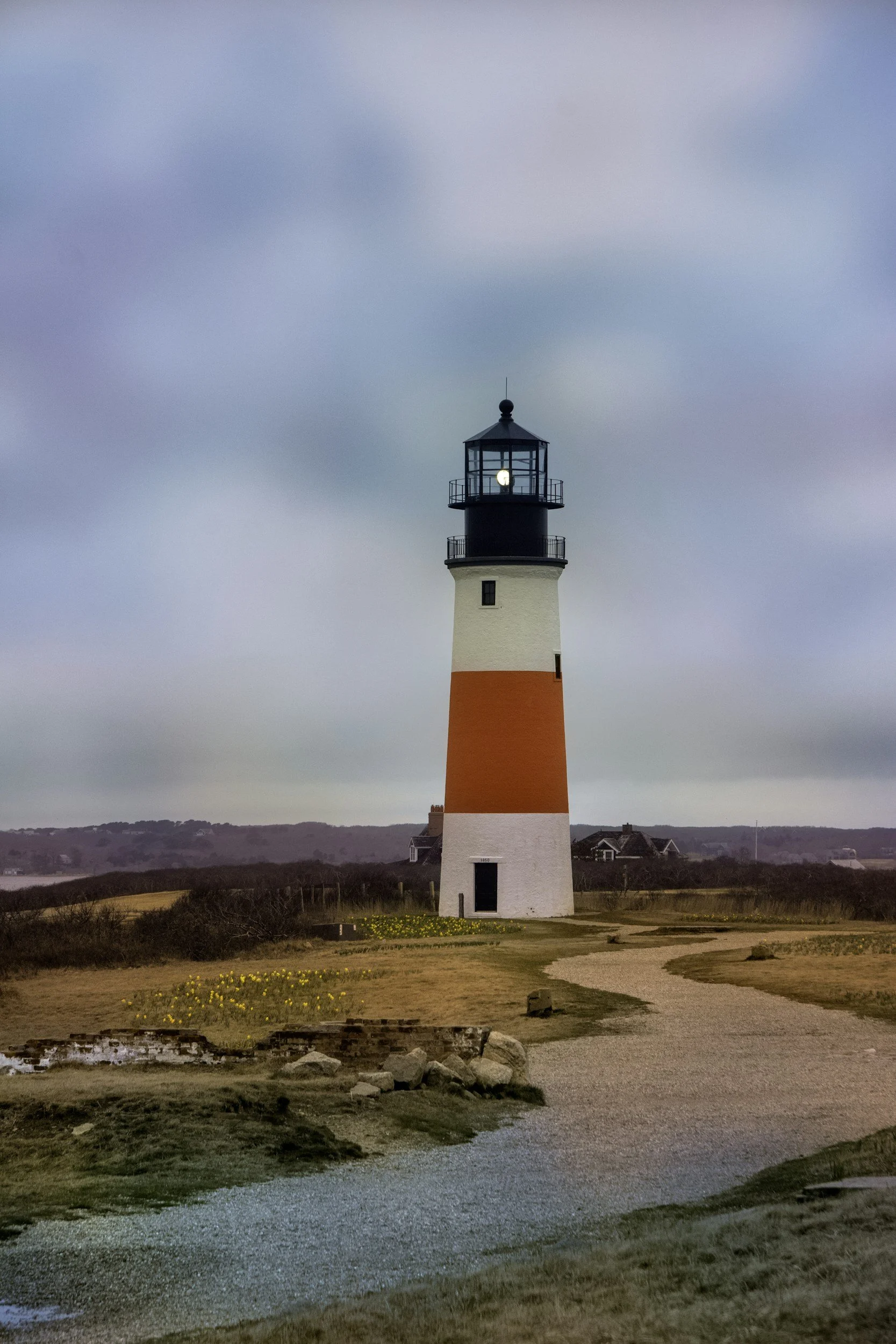 Lighthouse with a black top and white and orange banded tower, set against cloudy sky with landscape of grass and houses in the background.