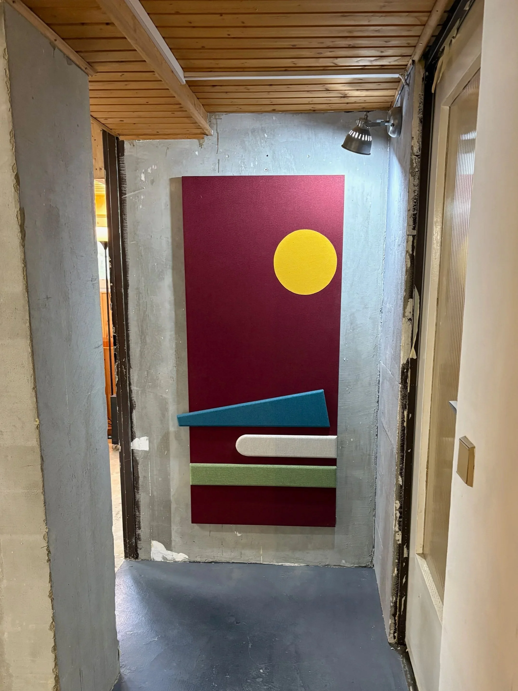 Ansicht eines Flurabschnitts mit Zierleisten und Wandpaneelen auf der rechten Seite, einem roten Wandpaneel mit gelbem Kreis, und farbigen geometrischen Holzformen auf dem Boden.