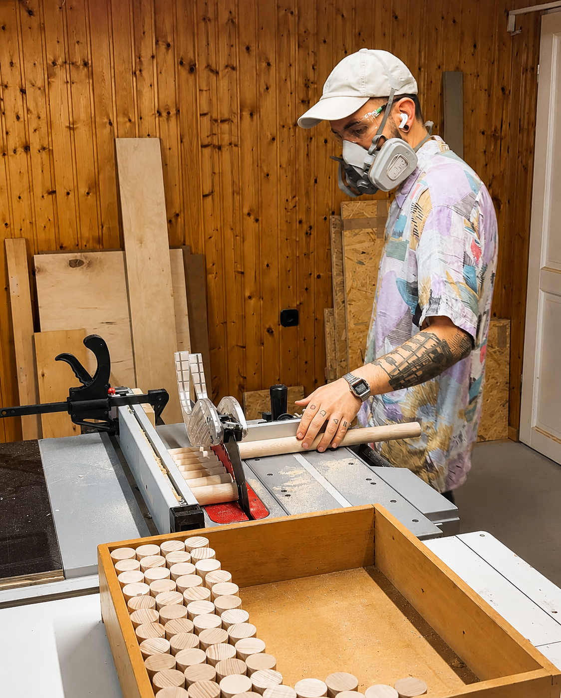 Junge Person arbeitet in einer Werkstatt mit Baumaterialien und einer Kreissäge, trägt eine Schutzmaske und Kopfhörer.