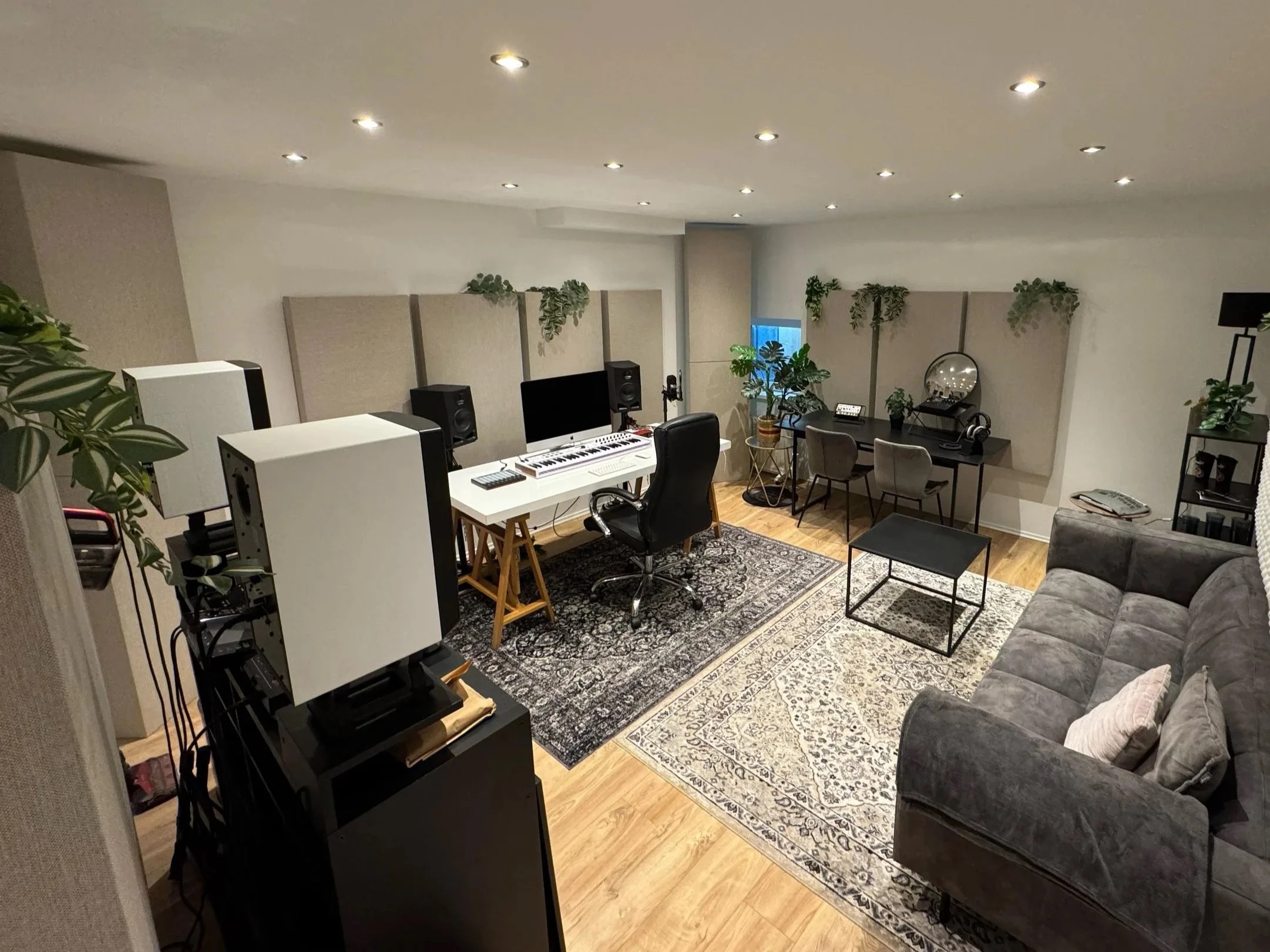 Ein Musikstudio mit einem weißen Keyboard, Monitor, Lautsprechern, mehreren Stühlen, einem Spiegel, zahlreichen Pflanzen und einem grauen Sofa auf einem orientalischen Teppich.