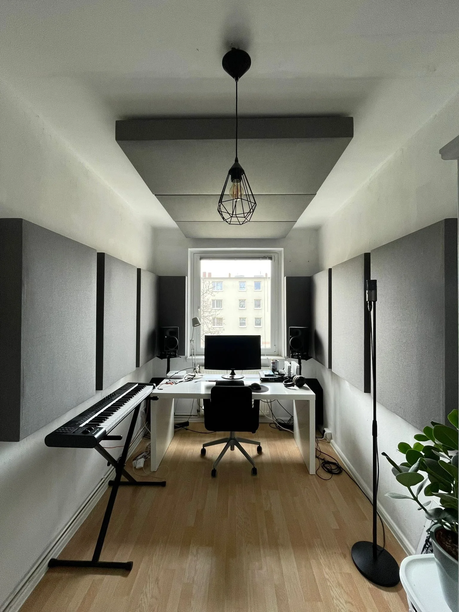 Musikproduktionsraum mit Arbeitsstation, Computermonitor, Lautsprechern, Keyboard, Mikrofon und akustischen Wandpaneelen, Blick auf ein Fenster.