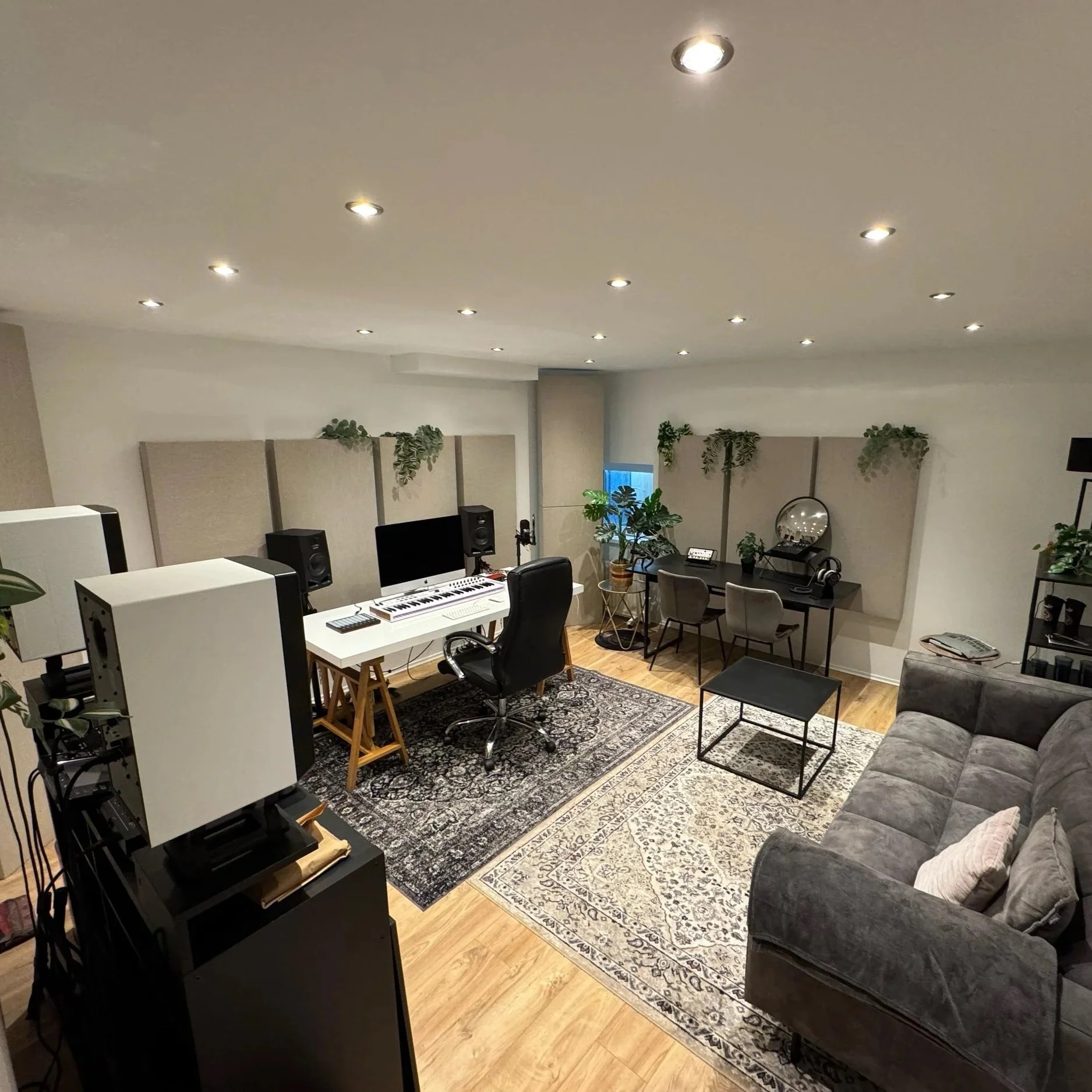 Ein Gästezimmer mit weißen Wänden, einem grauen Sofa, einem Teppich und mehreren Pflanzen, ausgestattet für Musikaufnahme oder Arbeit, mit einem Computer, Lautsprechern und Musikinstrumenten.