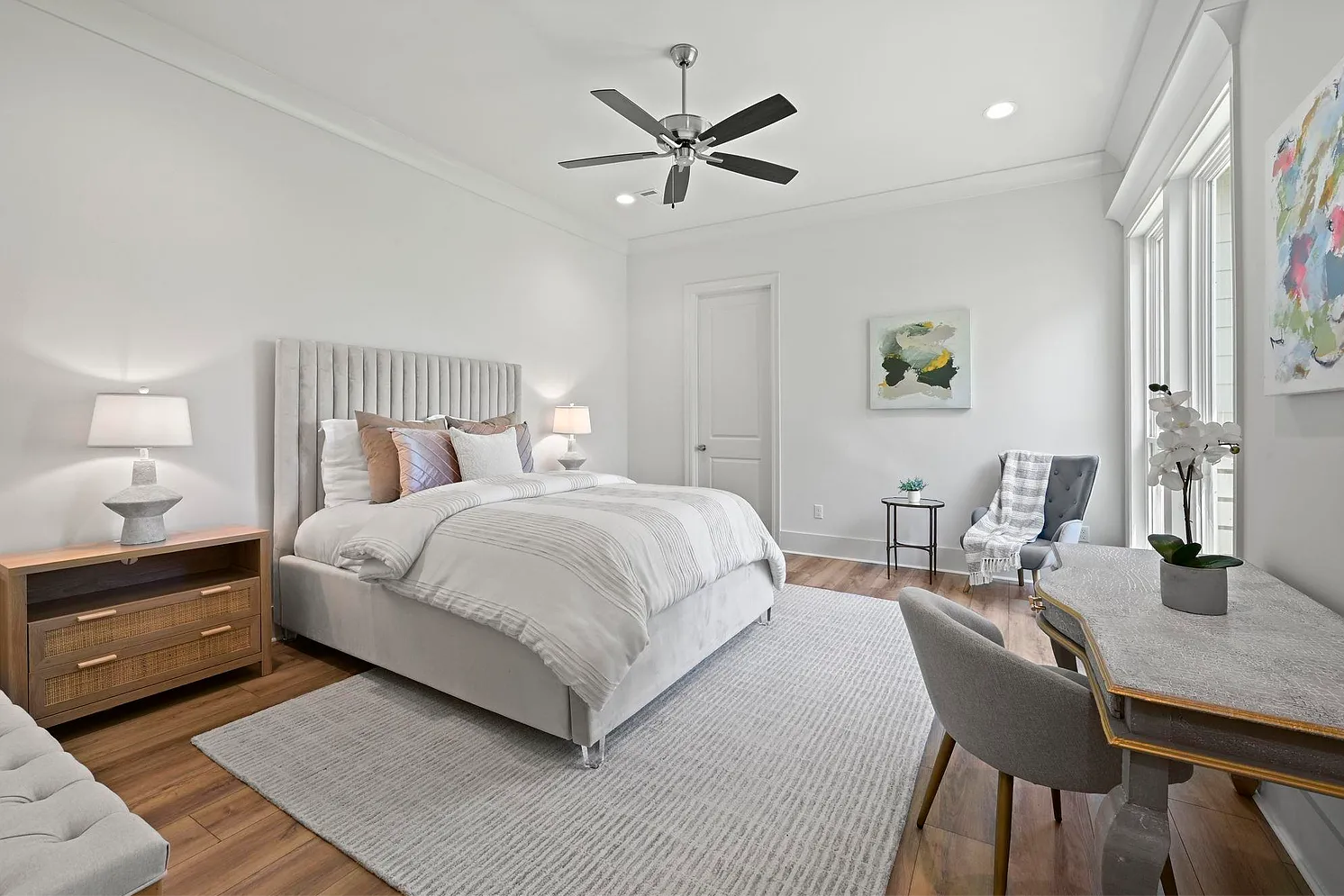 Bright bedroom with a large bed, wooden nightstands with lamps, a gray accent chair, decorative paintings, a console table with a flower pot, and a ceiling fan.
