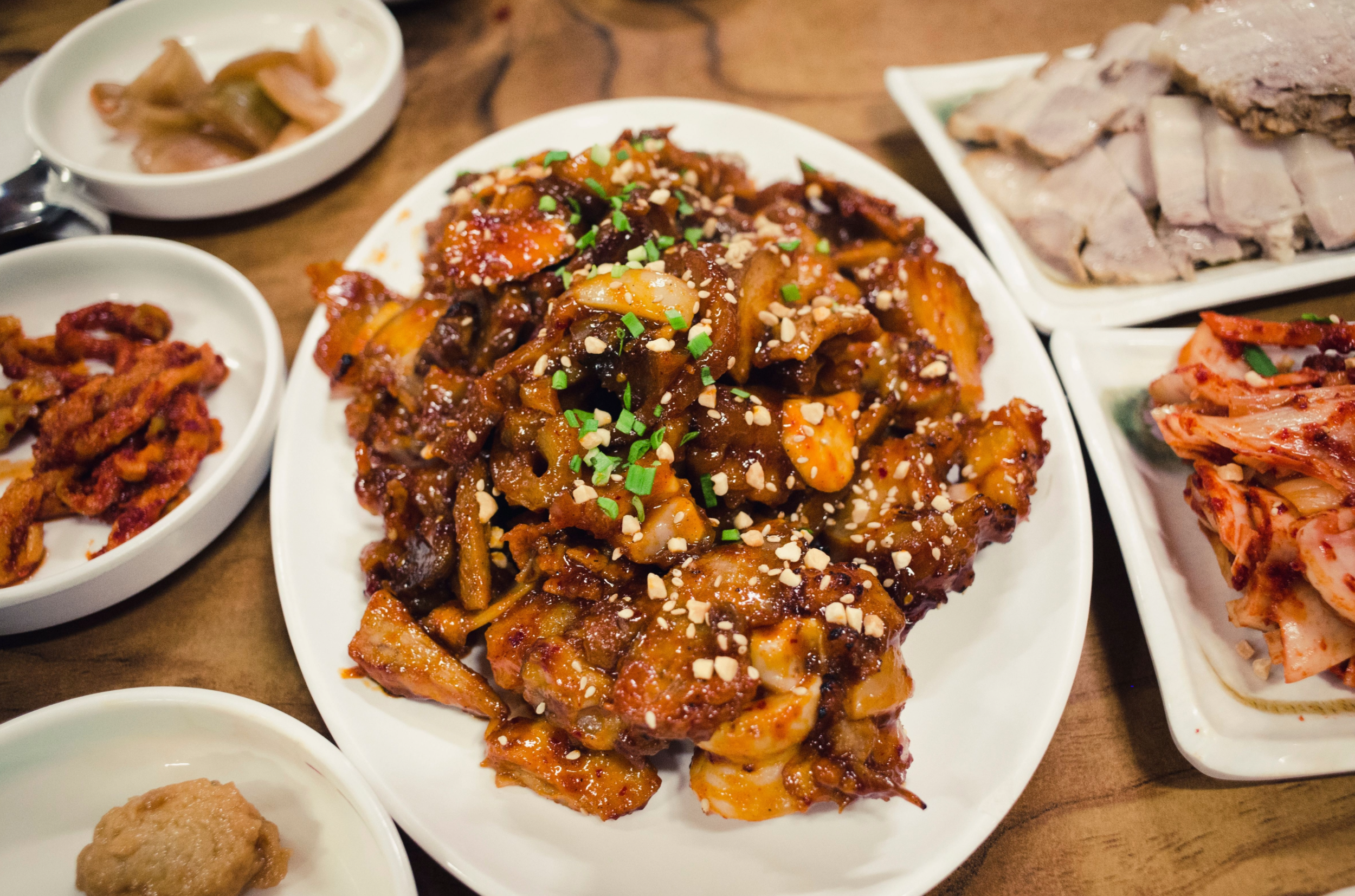 A variety of Korean dishes served on white plates, including spicy stir-fried eel with vegetables and chopped nuts, small side dishes with pickled vegetables, and a plate of sliced boiled pork.