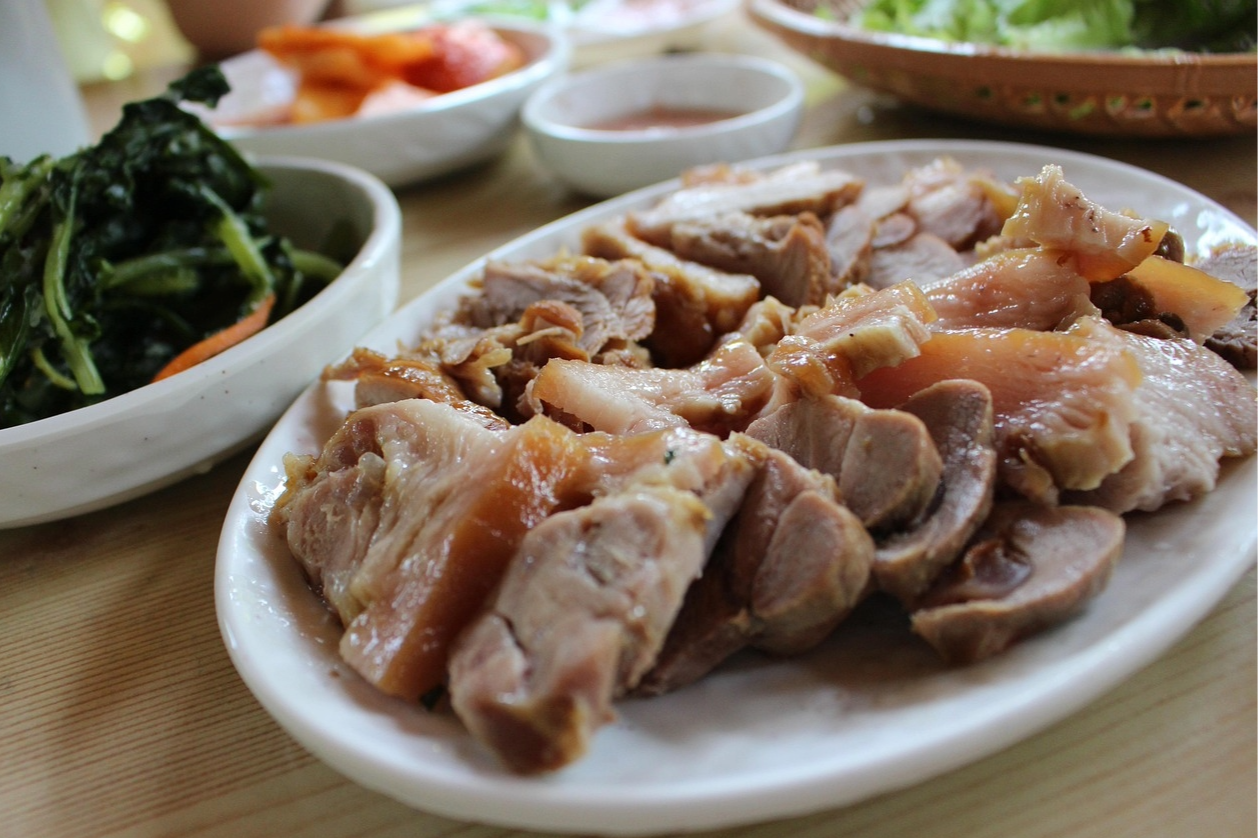 A plate of cooked chicken with some visible bones and skin, along with side dishes including leafy greens and dipping sauces, on a wooden table.