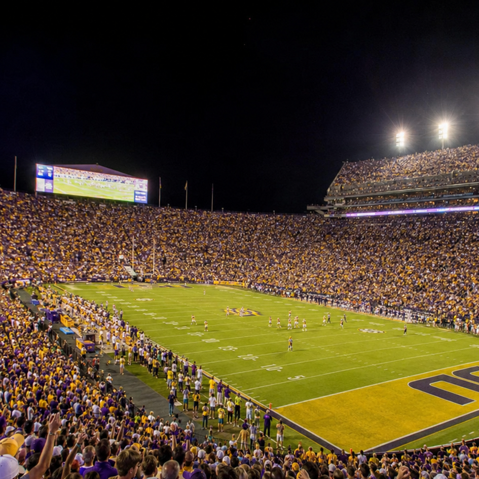 An American football stadium filled with spectators watching a game at night, with players on the field and bright lights above.