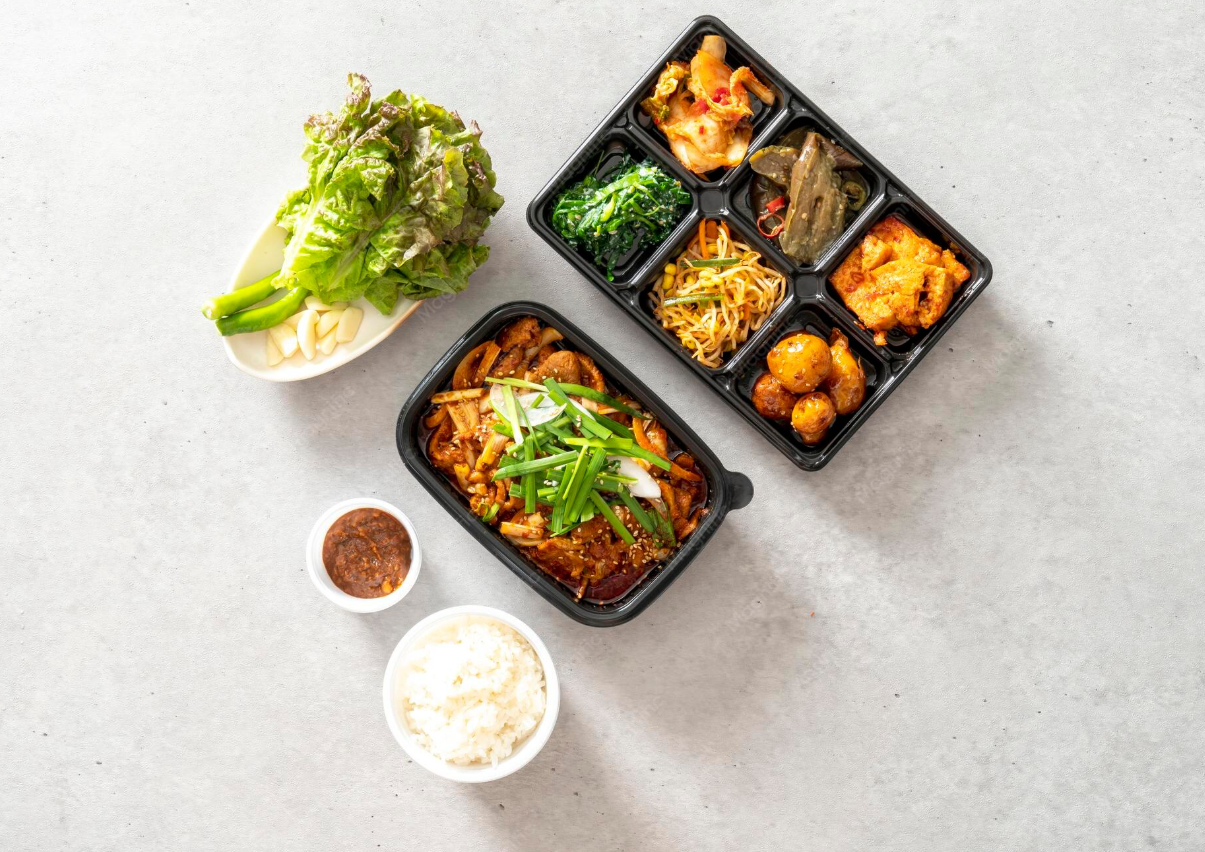 Korean meal with rice, soup, dipping sauce, and an assortment of side dishes in small containers, served with lettuce and garlic on the side.