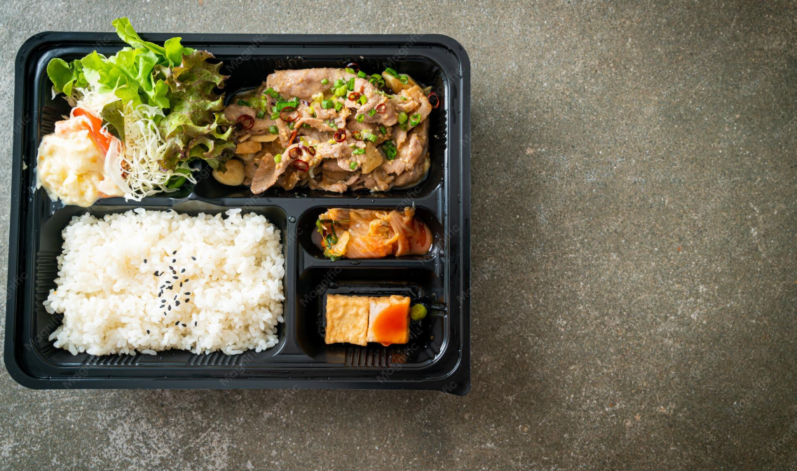 A black plastic tray with compartments filled with white rice topped with black sesame seeds, a small portion of lettuce salad with shredded cabbage and a tomato slice, cooked meat garnished with chopped green onions, a small serving of kimchi, and a slice of fried tofu with a small side of vegetables.