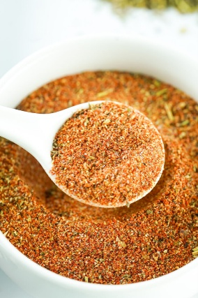A close-up of a white spoon filled with ground spice mixture, resting over a bowl of the same spice blend.