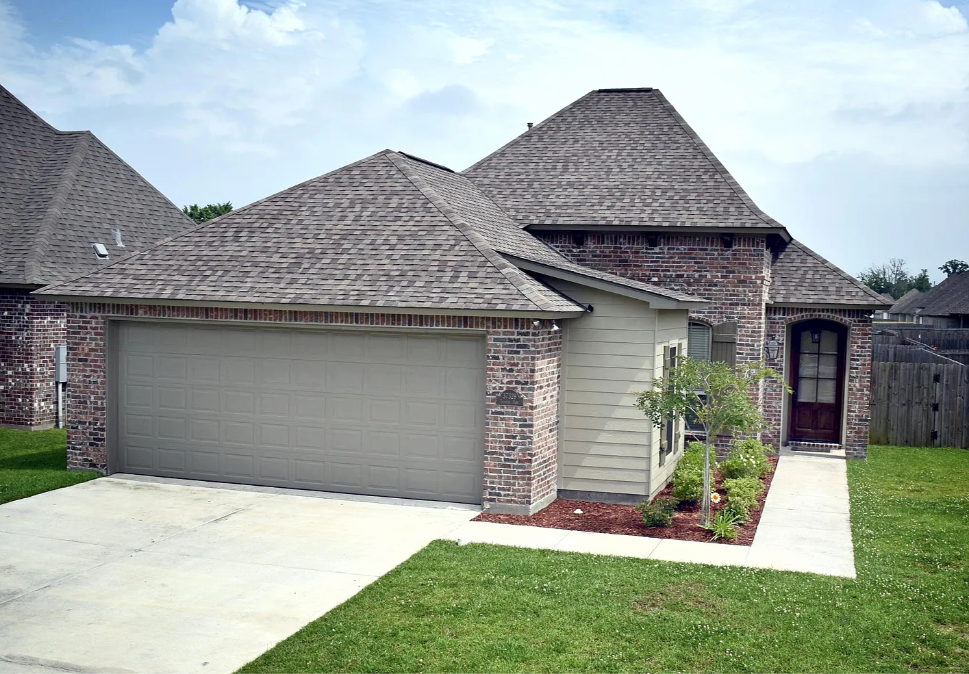 Front view of a brick and siding residential house with a concrete driveway and a small garden with a young tree and plants.