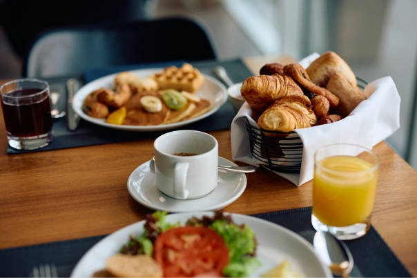 A breakfast setting with a plate of fried eggs, toast, and fruit, a basket of assorted pastries including croissants and bread, a cup of coffee, a glass of orange juice, and a glass of juice on a wooden table.